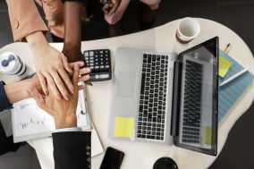 A diverse group of business professionals joining hands over a laptop, symbolizing collaboration and teamwork in a corporate setting.