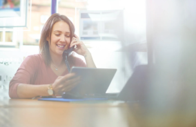 A woman seated at a desk, working on a laptop while holding a phone in her hand.